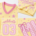 A close-up collage of a yellow athletic jersey with pink trim and pink printed details, including the word 'wildcats,' stars, bows, and large gingham-style numbers and letters.