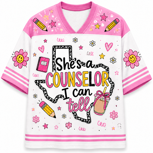 A white and pink V-neck graphic t-shirt featuring the text 'She's a COUNSELOR I can tell' inside a black-and-white outline map of Texas, decorated with school-themed doodles like pencils, flowers, stars, and hearts.