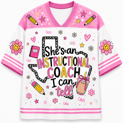 A girls' V-neck t-shirt with a white body and pink sleeves/collar, decorated with colorful cartoon graphics including smiley-face flowers, pencils, stars, and the text 'She's an INSTRUCTIONAL COACH I can tell'.