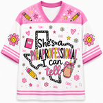 A women's short-sleeve V-neck t-shirt with a white body, pink collar and sleeve cuffs, decorated with a pink and black graphic that reads 'She's a PARaprofessional I can tell' along with doodles of pencils, flowers, and a drink.