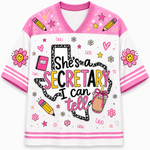 A white and pink short-sleeve V-neck t-shirt with a cartoon graphic that reads 'She's a SECRETARY I can tell'. The shirt features pink shoulder panels, striped sleeve cuffs, and various school-themed doodles like pencils, stars, flowers, and a coffee cup on a white background.