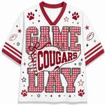 A white short-sleeve V-neck athletic-style shirt with red trim and the words 'GAME COUGARS DAY' in large red-and-white gingham block letters. The design includes paw prints, stars, footballs, and red stripes on the sleeves.