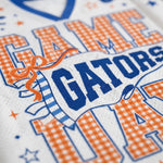 Close-up of a white athletic jersey with orange and blue printed lettering reading 'GAME GATORS' and '114', plus small star and ribbon graphics.