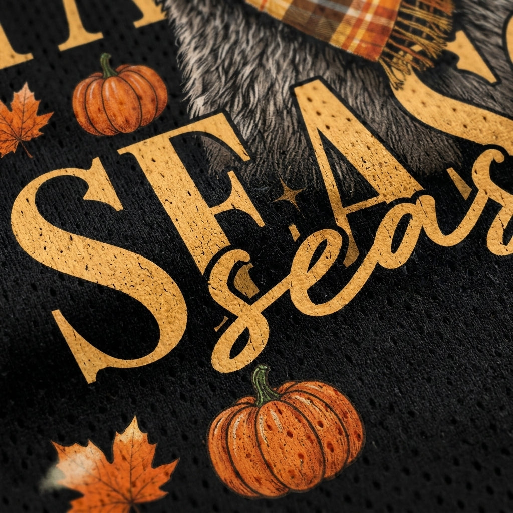 Close-up of a black knit fabric with a fall-themed graphic print featuring the words 'SEASONS', orange pumpkins, autumn leaves, and part of a plaid scarf.