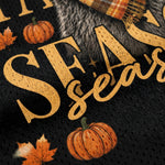 Close-up of a black knit fabric with a fall-themed graphic print featuring the words 'SEASONS', orange pumpkins, autumn leaves, and part of a plaid scarf.