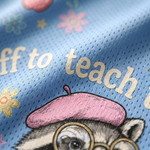 Close-up of a light blue mesh fabric printed with the phrase 'off to teach' in cream lettering, decorated with small pastel flowers and a cartoon raccoon wearing a pink beret and round glasses.