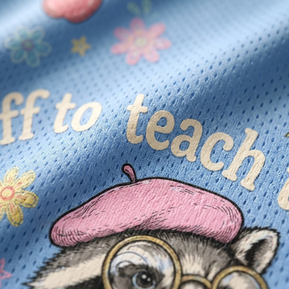 Close-up of a light blue mesh fabric printed with the phrase 'off to teach' in cream lettering, decorated with small pastel flowers and a cartoon raccoon wearing a pink beret and round glasses.