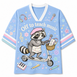A light blue short-sleeve V-neck t-shirt featuring a cartoon raccoon riding a scooter with musical notes and school supplies. The graphic includes the text 'Off to teach music' and is decorated with musical instruments, stars, and flowers on the sleeves.