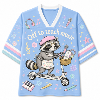 A light blue short-sleeve V-neck t-shirt featuring a cartoon raccoon riding a scooter with musical notes and school supplies. The graphic includes the text 'Off to teach music' and is decorated with musical instruments, stars, and flowers on the sleeves.