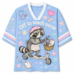 A light blue V-neck short-sleeve T-shirt with a cartoon raccoon riding a scooter on the front, wearing a pink beret and nurse apron. The shirt has the text 'Off to teach nurse' and is decorated with pastel pink, purple, and green medical-themed doodles including a stethoscope, heart monitor, bandage, stars, and flowers.