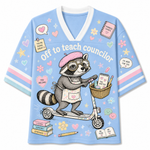 A light blue V-neck t-shirt with a cartoon raccoon riding a scooter, featuring the text 'Off to teach councilor' and various cute school-themed illustrations like books, hearts, and stars.