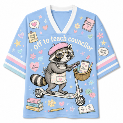A light blue V-neck t-shirt with a cartoon raccoon riding a scooter, featuring the text 'Off to teach councilor' and various cute school-themed illustrations like books, hearts, and stars.