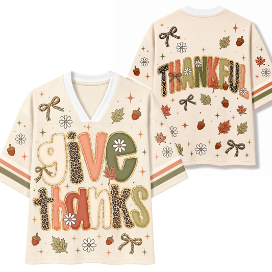 A cream-colored short-sleeve V-neck t-shirt shown from both front and back. The front features the words 'give thanks' in a playful, textured lettering style with leopard print, orange, and olive green tones, decorated with autumn leaves, acorns, bows, and daisies.