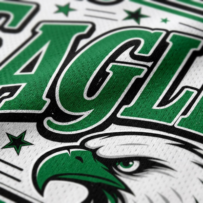 Close-up of a white athletic jersey or fabric graphic featuring large green 'EAGLE' lettering outlined in black, green stars, and part of a green eagle head illustration.
