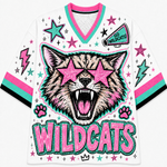 A white V-neck athletic jersey with a colorful graphic of a wildcat wearing pink star-shaped sunglasses, surrounded by stars, lightning bolts, paw prints, and the word 'WILDCATS' in large pink letters. The jersey has pink and teal trim on the collar and sleeve cuffs, with black stripes on the sleeves.