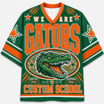 A green and orange V-neck jersey-style graphic top with the words 'WE ARE GATORS' and a large alligator illustration on the front, plus 'CUSTOM SCHOOL' near the hem. The design includes orange lettering, white stars, and striped sleeve accents on a plain light background.