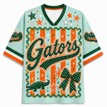 A light mint green V-neck t-shirt featuring Florida Gators graphics, including the word 'Gators' in green with white dots, orange stars, an alligator, and a bow on a striped background.