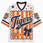 A short-sleeve V-neck jersey-style shirt with a tiger-themed graphic. The main body is off-white, with bold orange and navy blue graphics including the word 'Tigers' in script, stars, paw print, bow, stripes, and tiger head illustrations on the sleeves.