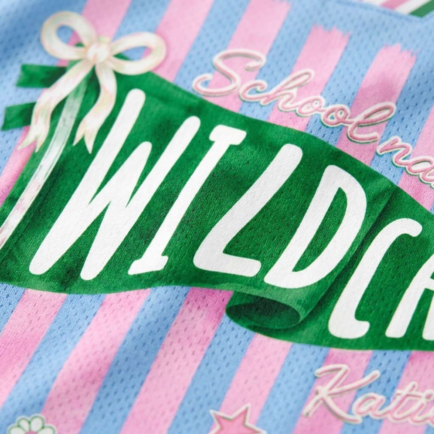 Close-up of a green graphic banner reading 'WILDCATS' in white letters, layered over a pink and light blue striped mesh fabric with white script text and a cream ribbon illustration.