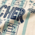 Close-up of a cream-colored fabric item, likely a towel or blanket, featuring navy blue text that reads 'TEACHER' and decorative illustrations in a light blue and navy design.