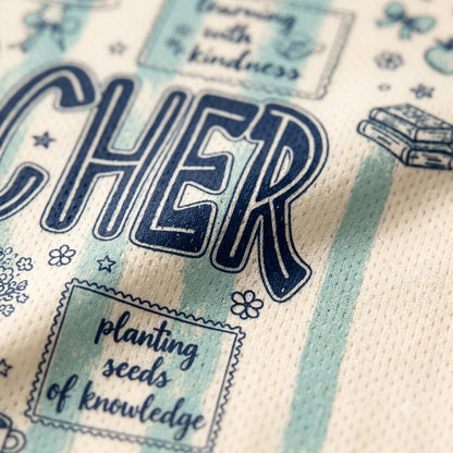 Close-up of a cream-colored fabric item, likely a towel or blanket, featuring navy blue text that reads 'TEACHER' and decorative illustrations in a light blue and navy design.