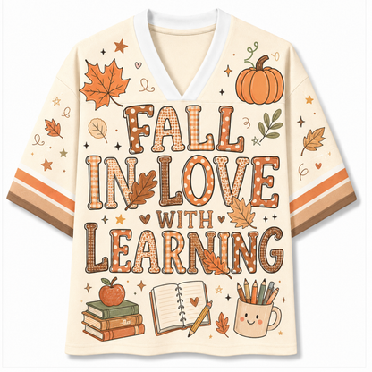 A cream-colored V-neck t-shirt with a cozy autumn theme, featuring the text 'FALL IN LOVE WITH LEARNING' in a plaid pattern, decorated with illustrations of pumpkins, maple leaves, books, and a smiling pencil cup.