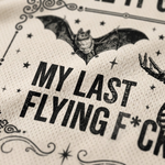 Close-up of a cream-colored textured fabric printed with a black bat illustration, stars, and the phrase 'MY LAST FLYING F*CK' in distressed bold lettering.