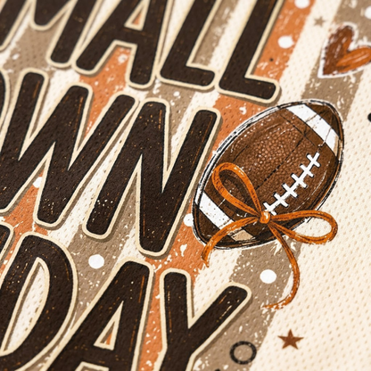 Close-up of a decorative printed design featuring large dark brown letters, an illustrated brown football with white laces, and an orange ribbon bow on a textured cream background.