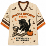 A beige V-neck t-shirt with a Halloween-themed graphic print. The design features a black kitten kneading a ghost biscuit on a cutting board, surrounded by a pumpkin, a bowl of dough, and stars.