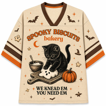 A beige V-neck t-shirt with a Halloween-themed graphic print. The design features a black kitten kneading a ghost biscuit on a cutting board, surrounded by a pumpkin, a bowl of dough, and stars.
