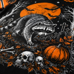 A close-up of a Halloween-themed graphic print featuring a raccoon, pumpkins, skulls, bats, crosses, and an orange full moon on a black background.