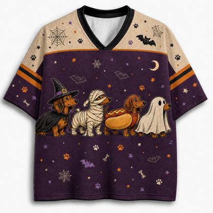 A short-sleeve V-neck t-shirt featuring a Halloween-themed graphic print with four dachshunds dressed as a witch, mummy, hot dog, and ghost, set against a purple background with spiderwebs, bats, and stars. The upper chest and shoulders feature a beige patterned print with spiderwebs and paw prints.