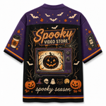 A purple short-sleeve t-shirt with an all-over Halloween-themed graphic print on the back. The design features a large retro TV showing a jack-o'-lantern, 'Spooky Video Store' text, pumpkins, bats, skulls, ghosts, and 'spooky season' script.