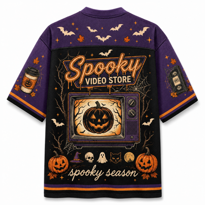 A purple short-sleeve t-shirt with an all-over Halloween-themed graphic print on the back. The design features a large retro TV showing a jack-o'-lantern, 'Spooky Video Store' text, pumpkins, bats, skulls, ghosts, and 'spooky season' script.