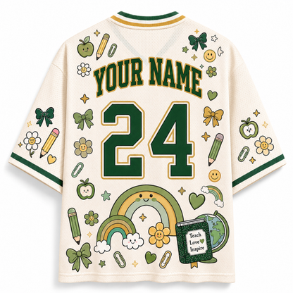 A cream-colored short-sleeve jersey-style shirt shown from the back, featuring green and gold trim, customizable 'YOUR NAME' text, and the number '24' in large green block letters. The shirt is decorated with school-themed illustrations including rainbows, pencils, flowers, bows, paperclips, smiley faces, and a book that reads 'Teach Love to Inspire'.