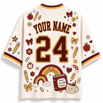 A cream-colored short-sleeve jersey shown from the back, featuring customizable text 'YOUR NAME' and the number '24' in dark brown with gold outlines. The shirt is decorated with various school-themed illustrations including rainbows, pencils, apples, stars, and a book that reads 'Teach Love Inspire'.