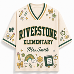 A cream-colored short-sleeve V-neck jersey-style shirt with green and gold trim, featuring 'RIVERSTONE ELEMENTARY' and 'Mrs. Smith' printed on the front, surrounded by school-themed illustrations like pencils, flowers, stars, and a rainbow.