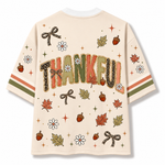 A cream-colored short-sleeve t-shirt with a colorful autumn-themed graphic print. The front features the word 'THANKFUL' in large, textured letters filled with leopard print, stripes, and leaf patterns, surrounded by maple leaves, acorns, bows, and daisies.