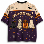 A short-sleeve graphic T-shirt shown from the back, featuring a Halloween-themed design with the text 'THE SPOOKY SQUAD' above four dog characters dressed as a witch, mummy, hot dog, and ghost. The shirt has a beige upper section and a dark purple lower section with orange, black, and white graphic details.