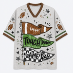A short-sleeve V-neck athletic-style shirt with a white mesh-like base and brown trim, decorated with football motifs and the words 'TOUCH DOWN GAME DAY' across two pennants. The shirt is shown flat against a light gray background.