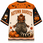 A short-sleeve crewneck shirt shown from the back, featuring a colorful autumn-themed graphic print with a raccoon in a beanie and scarf, pumpkins, leaves, bats, paw prints, and the text 'AUTUMN BANDITS' on a cream background with orange and black sleeves.