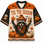 A festive Halloween-themed V-neck jersey-style shirt featuring a raccoon wearing a knit hat and scarf, surrounded by pumpkins, autumn leaves, bats, paw prints, and the text 'TIS THE SEASON'. The sleeves and shoulders are orange and black, with printed graphics of a coffee cup and a jack-o'-lantern.