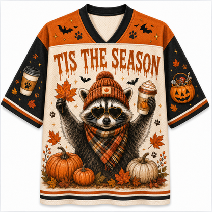 A festive Halloween-themed V-neck jersey-style shirt featuring a raccoon wearing a knit hat and scarf, surrounded by pumpkins, autumn leaves, bats, paw prints, and the text 'TIS THE SEASON'. The sleeves and shoulders are orange and black, with printed graphics of a coffee cup and a jack-o'-lantern.