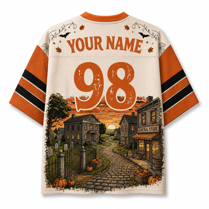 A short-sleeve football-style jersey shown from the back, with orange and black sleeve stripes, a cream body, and large orange text reading 'YOUR NAME 98' above a Halloween village scene. No real person or environment is present; this is a clean product rendering on a white background.