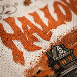 Close-up of a Halloween-themed textile design featuring large orange distressed lettering and a black-and-orange haunted house illustration on a textured cream fabric.