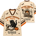 A cream-colored V-neck t-shirt with Halloween-themed graphics, shown in both front and back views. The design features a black cat kneading a biscuit, a pumpkin, bats, stars, and the text 'SPOOKY BISCUITS bakery' and 'WE KNEAD EM YOU NEED EM'.