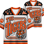 Two views of a custom V-neck athletic jersey featuring 'THS TIGERS' in orange lettering, a roaring tiger graphic, paw prints, and orange and black accents on a white background.