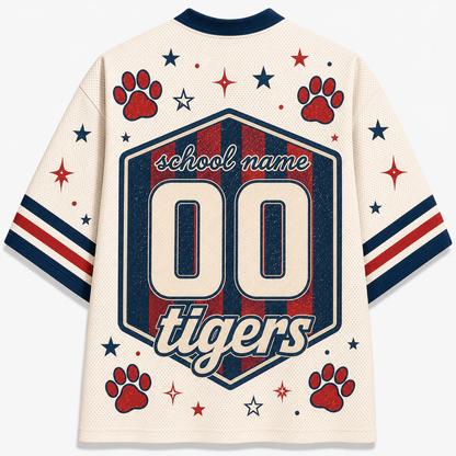 A cream-colored mesh short-sleeve jersey shown from the back, featuring a large navy and red hexagonal graphic with the text 'school name', '00', and 'tigers'. The design is decorated with red paw prints, red and navy stars, and red and navy stripes on the sleeves.
