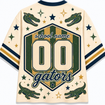 A cream-colored athletic jersey shown from the back, featuring green and navy blue graphics. The design includes two alligator heads near the shoulders, green and gold stars, a central shield with the text 'school name', '00', and 'gators', and two alligators near the lower hem.