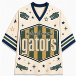 A cream-colored custom sports jersey featuring a V-neck, navy blue trim, and green and gold graphics. The front displays a large hexagonal shield with the text 'school name gators' in a retro font, flanked by green alligator illustrations and gold and navy blue stars.